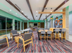 Modern lounge area with wooden chairs, tables, and a colorful carpet. Walls have green accents and a large window, creating an open and bright space.