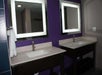 Two bathroom sinks with rectangular mirrors featuring built-in lighting, set against a purple wall. White towels hang on a ring next to the left sink.