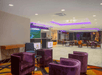 A modern hotel lobby with purple armchairs, small tables, a reception desk, and ceiling lights with purple accents.