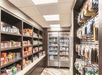 A large market area with brown shelves full of snacks on the left side, toiletries on the wall to the left, and refrigerators on the back wall.