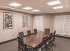 A conference room with two small windows with white curtains and a long dark brown table with office chairs around it.