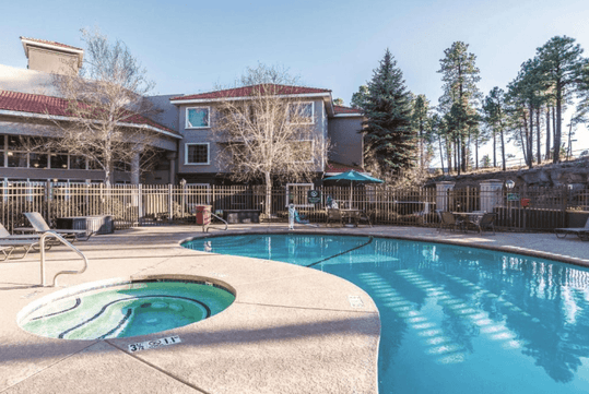 An our door swimming pool and small hot tub with lounge chairs and tables around it with bare trees and the La Quinta by Wyndham Flagstaff in the background.
