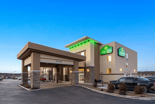 Exterior view of a La Quinta Inn & Suites hotel with two-story building, green signage, and entrance canopy. Surrounded by a parking area and small landscaped areas.