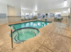 Indoor hotel pool area with a small circular hot tub in the foreground and a larger rectangular pool beyond. Several lounge chairs and a table with chairs are placed around the pool.