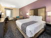 Hotel room with a large bed, patterned carpet, purple accent wall, desk, seating area, two lamps, and a window with patterned curtains.