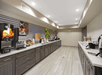 Hotel breakfast area with a long counter featuring coffee machines, juice dispensers, cereal, pastries, and fruit. Walls are decorated with food images. Bright overhead lighting.