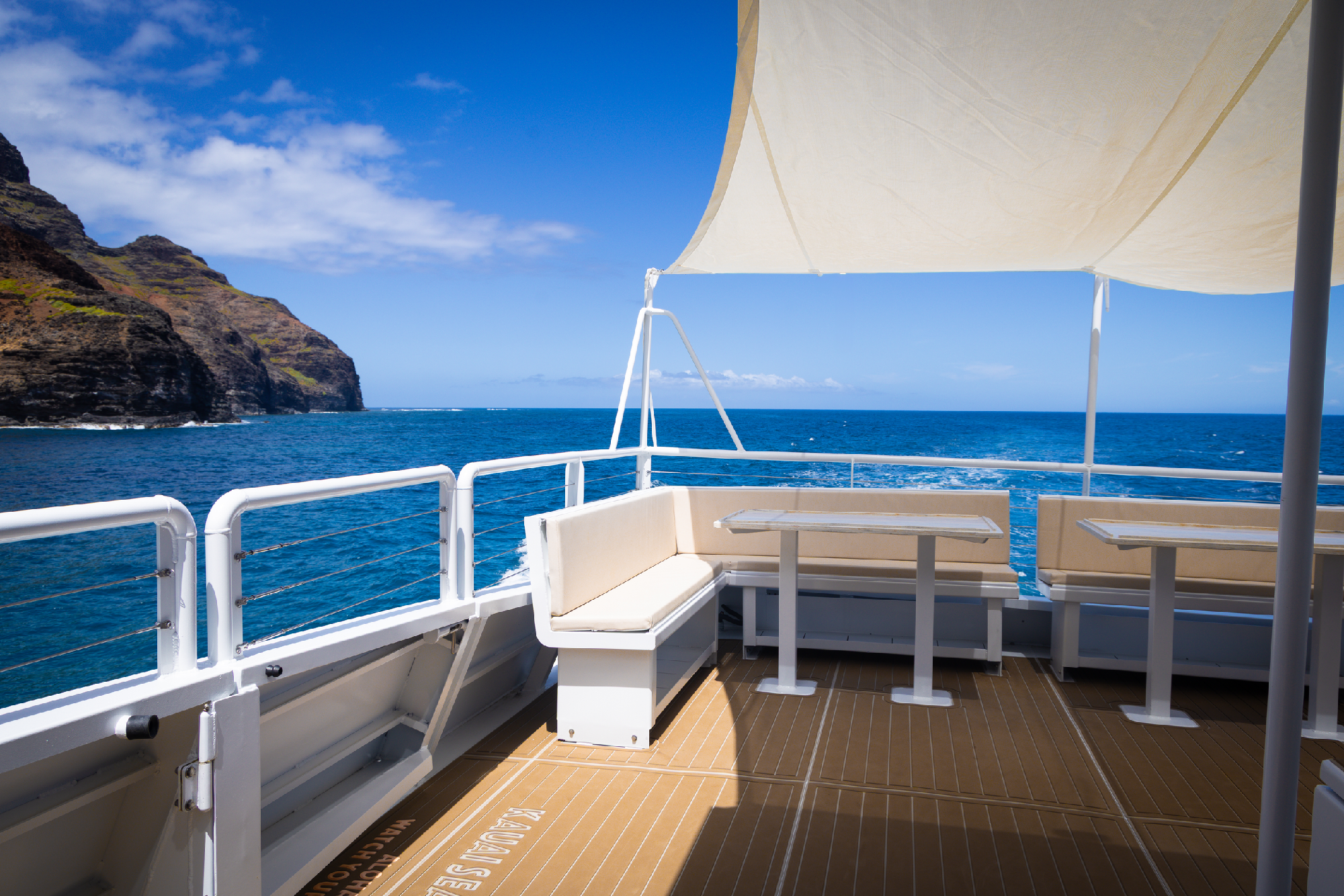 Open deck of a boat with benches and tables, shaded by a canopy, overlooking blue ocean water and rocky cliffs under a partly cloudy sky.