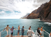 A group of people stand and talk on the deck of a boat near a rocky coastline with blue water and cliffs under a partly cloudy sky.