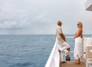 A man and a woman stand on the deck of a boat, looking at each other, with the ocean and a cloudy sky in the background.