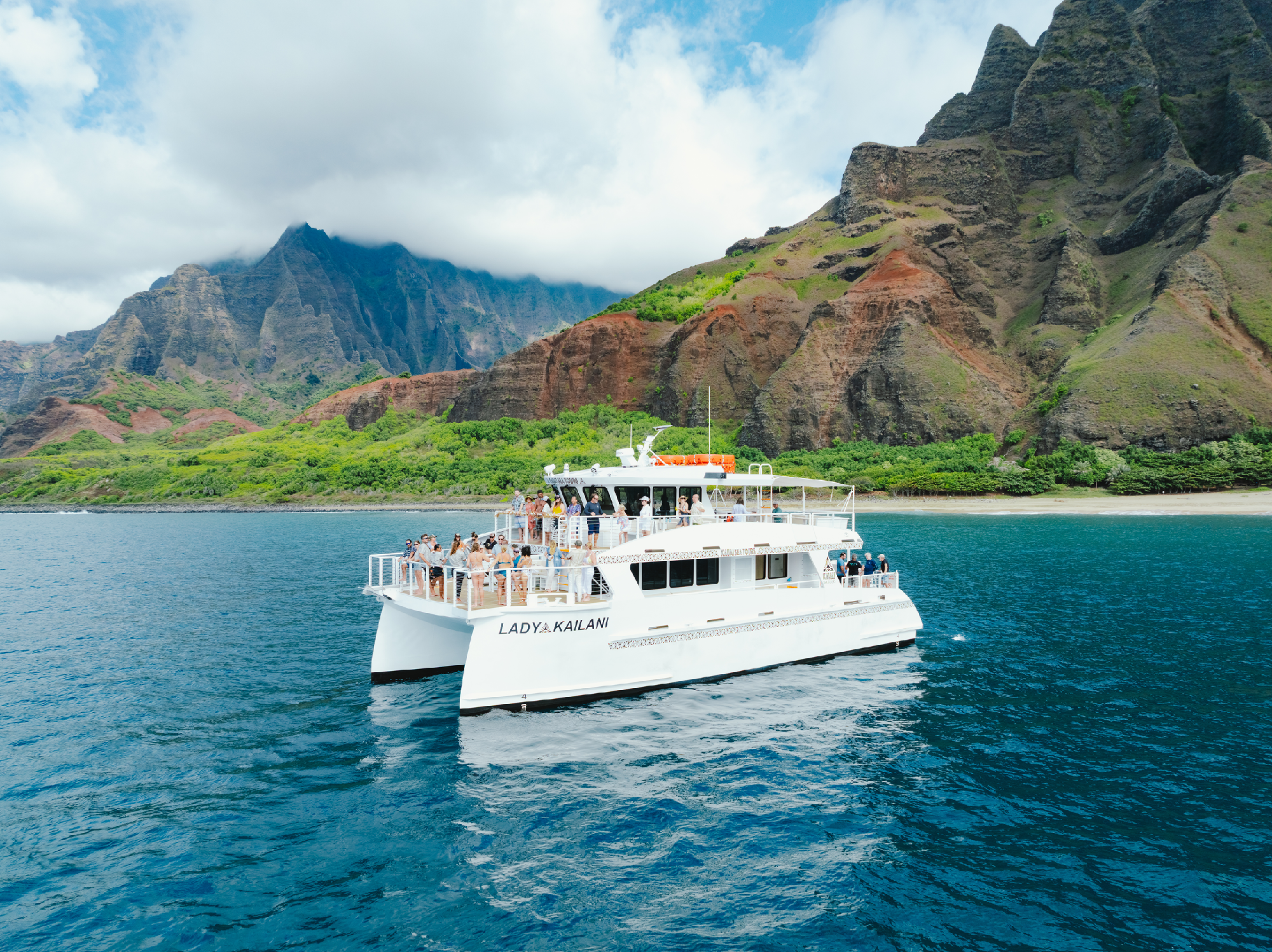 A white catamaran with people on board floats on blue water near a scenic mountainous coastline with green and red cliffs under a partly cloudy sky.
