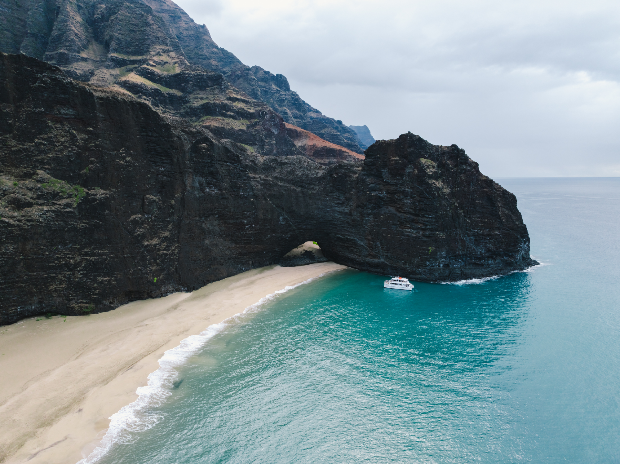 A white boat is anchored near a large rock arch on a sandy beach with steep, rugged cliffs by the ocean under a cloudy sky.