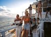 A group of people enjoy a boat ride on the ocean, some standing on deck and others seated, with sunlight and clouds in the background.