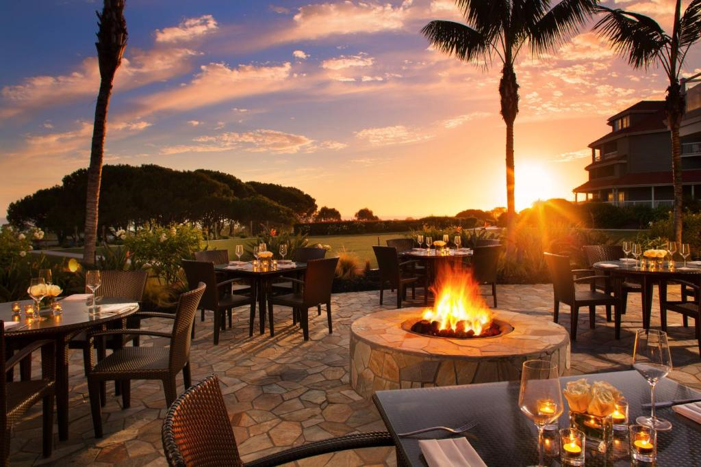 Outdoor patio with tables, chairs, and a central fire pit at sunset. Palm trees and a distant building are silhouetted against the sky.