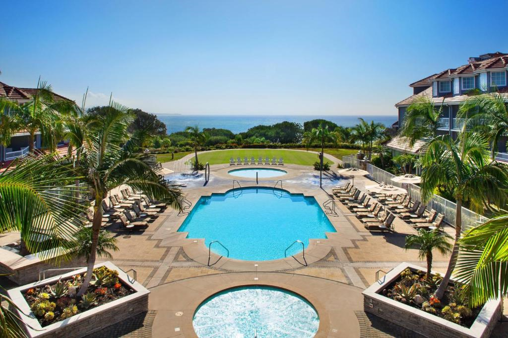 Aerial view of a resort with a large pool, surrounded by deck chairs, palm trees, and landscaped gardens, overlooking the ocean.