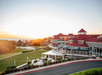 Resort with red-roofed buildings, patio umbrellas, and a green lawn, set against a sunset sky and distant ocean view.