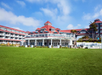 A large, multi-story building with red roofs and white trim sits behind a grassy lawn. The building features multiple windows and balconies under a blue sky with scattered clouds.