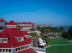 Red-roofed resort buildings overlooking lush green lawns and palm trees, with a clear blue sky and ocean view in the background.
