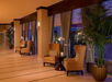 A hotel lobby with plush armchairs and small tables along a windowed wall. Potted plants and curtains add decor. Warm lighting creates an inviting atmosphere.