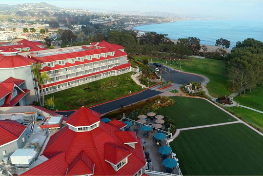 Aerial view of a coastal resort with red-roofed buildings, green lawns, and outdoor seating. The ocean is visible in the background, along with surrounding hills and a road.