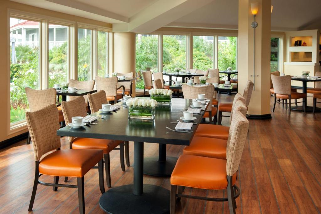 A sunlit dining area with wooden floors, large windows, and several tables set with white dishes and surrounded by wicker chairs with orange cushions.