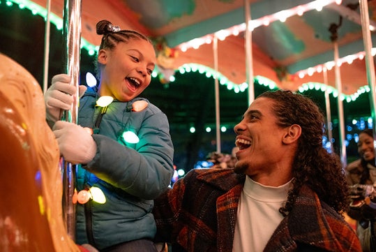 A man and a child with holiday lights ride a carousel, smiling joyfully.