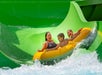 Three people are riding a yellow inflatable raft down a water slide, surrounded by splashing water and a bright green slide structure.