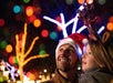 A man and a child wearing festive hats look up at colorful, blurred holiday lights in the night.