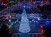 A large, brightly lit Christmas tree stands in front of an amusement park entrance at night, with roller coaster tracks and colorful holiday lights in the background.