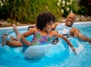 A child and an adult joyfully splash water while floating in swim tubes in a swimming pool on a sunny day.