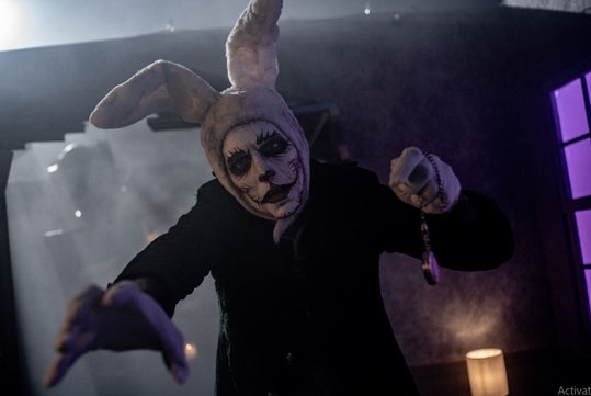 A sinister figure in a rabbit costume with exaggerated facial features and a menacing expression, reaching out towards the viewer in a dark, foggy setting.
