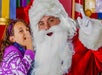 A child whispers to Santa Claus, who is wearing a red suit and white beard, in a festive setting with colorful decorations.