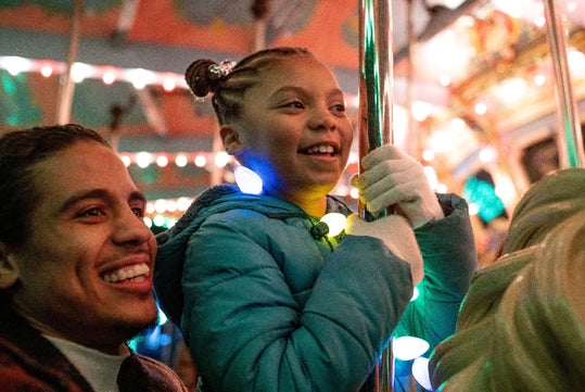 A young girl wearing gloves and a blue coat holds a pole on a carousel, smiling, with colorful lights and an adult smiling beside her.