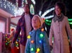 A child wearing a coat and colorful holiday lights around their neck walks outside at night with two adults; festive lights decorate the background.