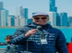 An older man in a Vendella jacket and cap holds a microphone and smiles, standing outdoors with a city skyline and water in the background.