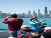 People on a boat take photos of a city skyline across blue water on a sunny day, with tall skyscrapers visible in the background.