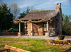 A rustic log cabin with a wooden porch and stone chimney, surrounded by trees. A green wagon sits beside it. A flower bed is in the foreground.