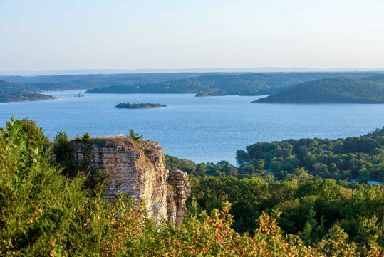 A scenic view of a wide lake with small islands, surrounded by green hills and a rocky cliff in the foreground under a clear sky.