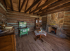 Rustic cabin interior with exposed wooden beams, a stone fireplace, a vintage green stove, and a round table with a tablecloth and fruit bowl.