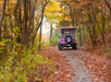 A pink jeep drives on a leaf-covered trail through a forest in autumn, surrounded by trees with colorful foliage.