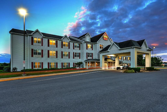A white, three-story hotel building with multiple windows, lit up at dusk. A car is parked under the entrance canopy. The sky is blue with pink clouds.