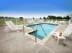 Outdoor swimming pool with metal handrails, surrounded by white lounge chairs and a fence. A drink and a book are placed on a chair. The sky is clear and blue.