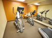 A small gym with cardio machines, including an elliptical, a stationary bike, and a treadmill, placed on a carpeted floor with a mirrored wall.