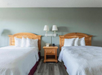 Two double beds with wooden headboards are in a hotel room. A nightstand with a lamp and a phone is between them. The walls are painted in a light green shade.