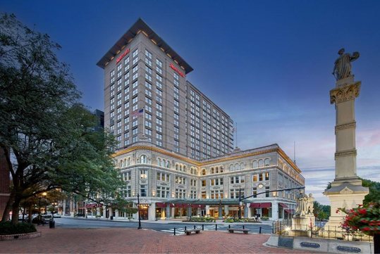 A large, multi-story hotel building on a city street corner with a statue in the foreground and trees nearby. The sky is a clear blue.