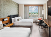 Hotel room with two double beds, a desk, chair, and a large window with a cityscape view. Walls are painted light with a patterned accent wall.
