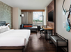 Modern hotel room with a large bed, chaise lounge, desk, TV, and hexagonal wall art. A window offers a view of the cityscape.