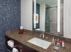 A bathroom with a granite countertop, a sink, toiletries, and a wall-mounted mirror. The shower area is visible in the reflection, featuring tiled walls and a glass door.