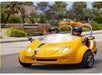 Two people in helmets smiling and sitting in a small yellow three-wheeled vehicle with a "GoCar" logo, driving on a sunny street.