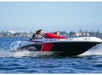 Two people wearing sunglasses are riding a speedboat on a sunny day, with the ocean and a distant shoreline in the background.