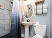 A bathroom with a pedestal sink, round mirror, toilet, folded towels on a shelf, and a shower with a gradient curtain and white tile walls.